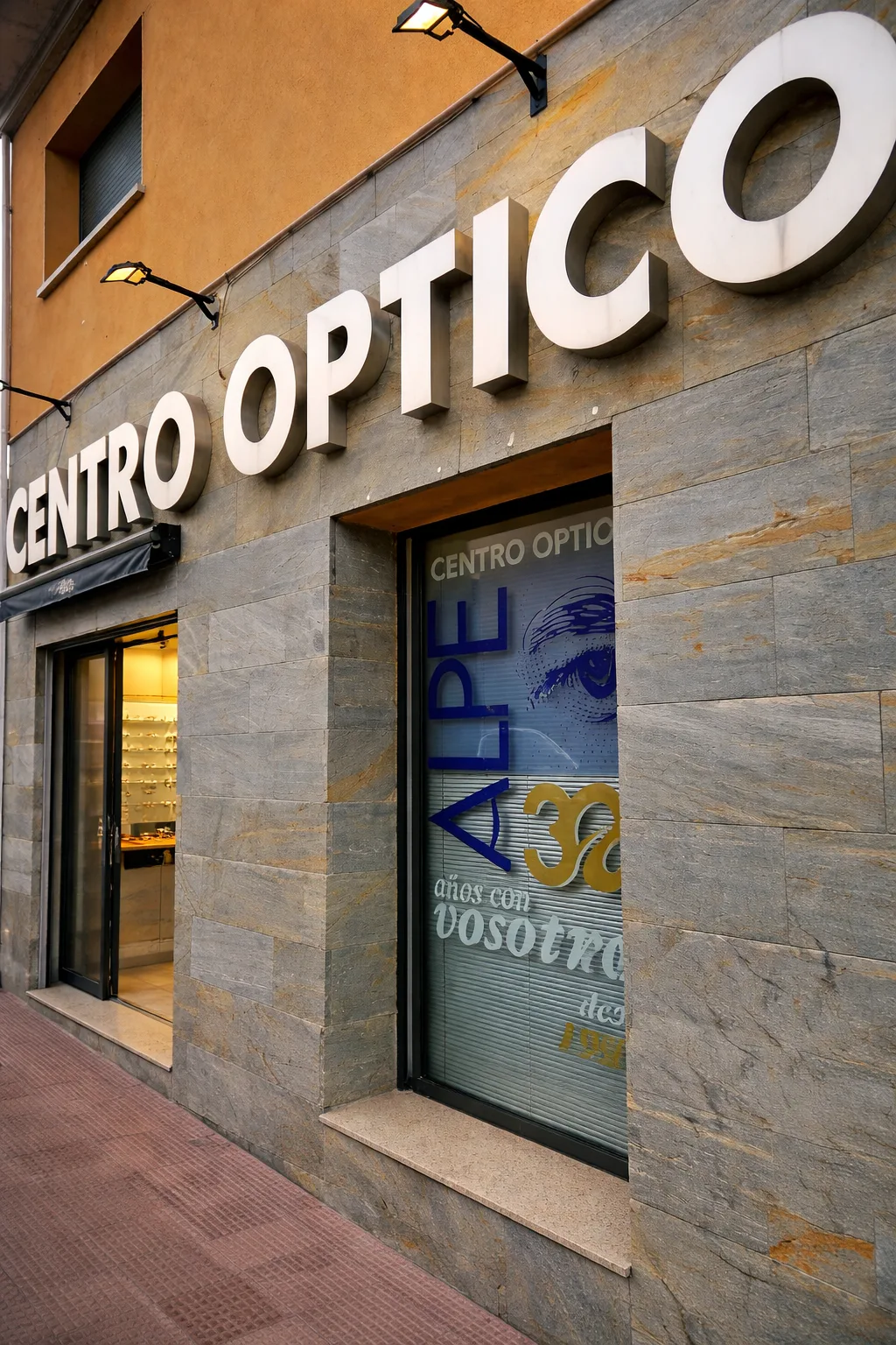 Fachada del Centro Óptico Alpe iluminada al anochecer, en Alpedrete